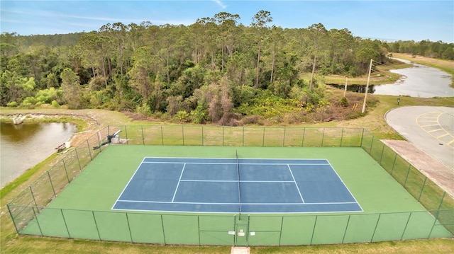 view of tennis court with a water view, fence, and a wooded view