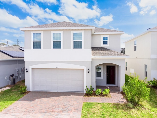 traditional home featuring stucco siding, decorative driveway, a garage, and roof with shingles