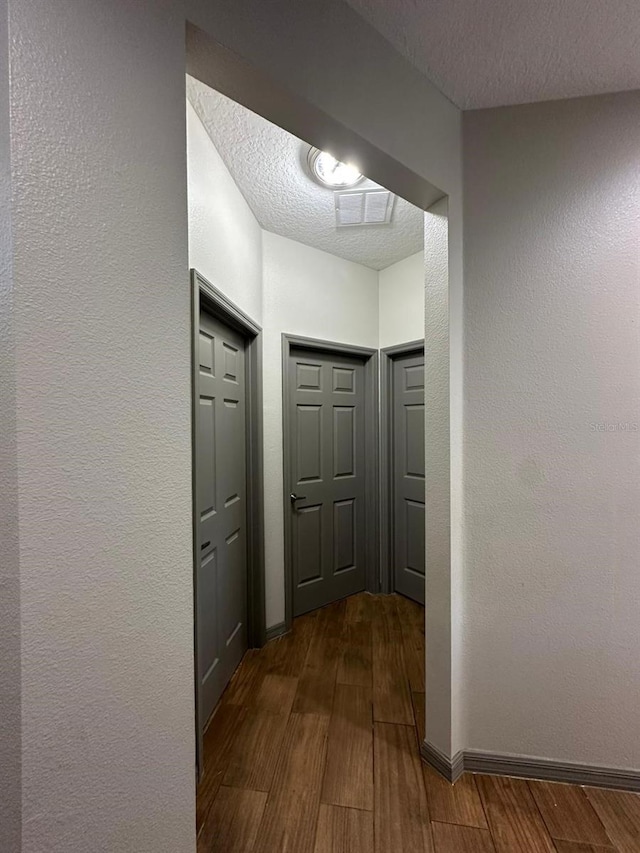 hall with a textured ceiling, a textured wall, dark wood-style flooring, visible vents, and baseboards