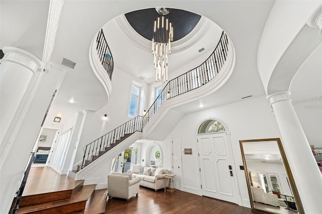 foyer entrance featuring stairway, a towering ceiling, dark wood-style floors, a notable chandelier, and ornate columns