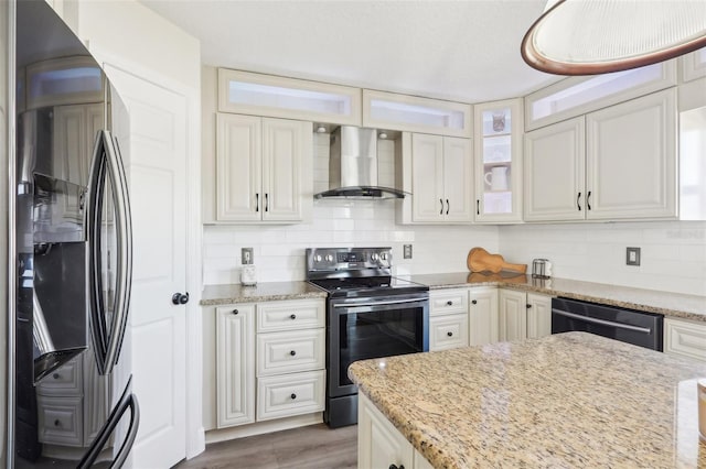 kitchen featuring glass insert cabinets, wall chimney range hood, light stone counters, decorative backsplash, and appliances with stainless steel finishes