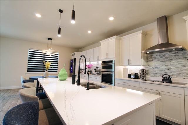 kitchen featuring backsplash, black electric stovetop, wall chimney range hood, double oven, and an island with sink