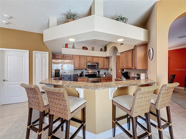 kitchen with brown cabinets, arched walkways, appliances with stainless steel finishes, a peninsula, and light tile patterned floors