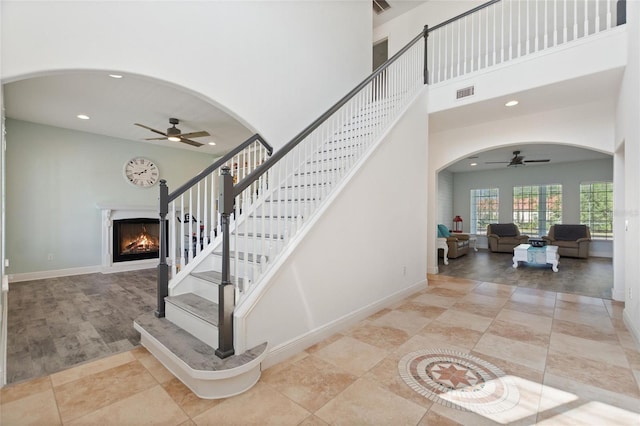 stairs with visible vents, a ceiling fan, arched walkways, and a warm lit fireplace