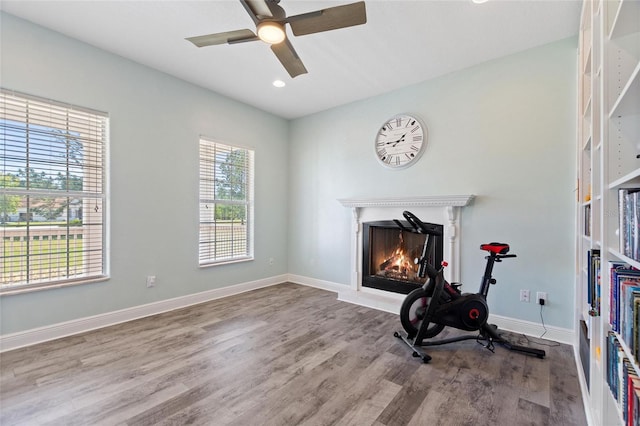 workout room featuring a lit fireplace, baseboards, a ceiling fan, and wood finished floors