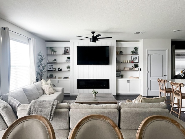 living room featuring visible vents, a textured ceiling, dark wood-style floors, a glass covered fireplace, and ceiling fan