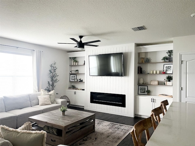 living area featuring visible vents, a textured ceiling, a glass covered fireplace, and dark wood-style flooring