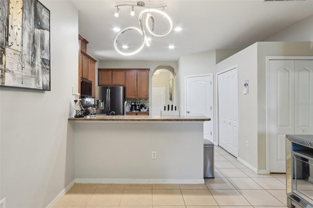 kitchen with brown cabinetry, light tile patterned floors, a peninsula, arched walkways, and stainless steel fridge