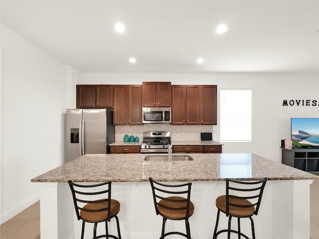 kitchen with light stone countertops, a kitchen island with sink, a sink, appliances with stainless steel finishes, and tasteful backsplash