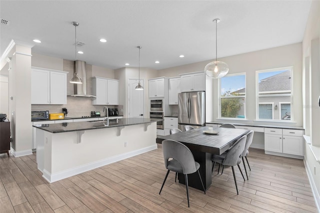 kitchen with a sink, wall chimney range hood, decorative backsplash, stainless steel appliances, and a kitchen island with sink