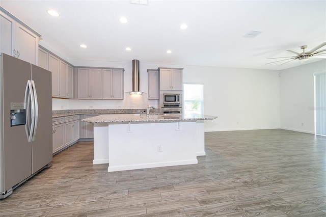 kitchen with stainless steel appliances, a center island with sink, gray cabinetry, wall chimney range hood, and a kitchen breakfast bar
