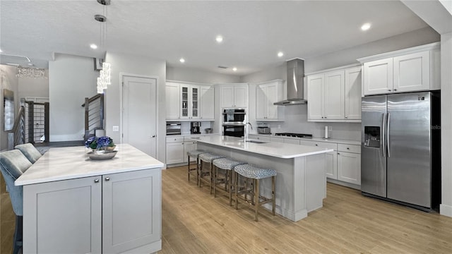kitchen featuring light wood-type flooring, stainless steel appliances, an island with sink, and wall chimney exhaust hood
