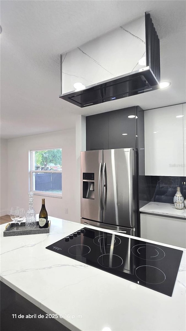 kitchen with stove, range hood, light stone countertops, stainless steel fridge, and white cabinetry