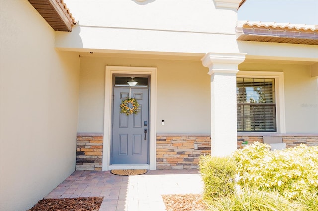 view of exterior entry with covered porch, a tiled roof, stone siding, and stucco siding