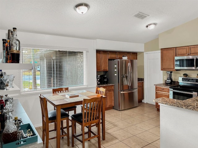 kitchen featuring brown cabinetry, a textured ceiling, appliances with stainless steel finishes, light tile patterned floors, and stone countertops