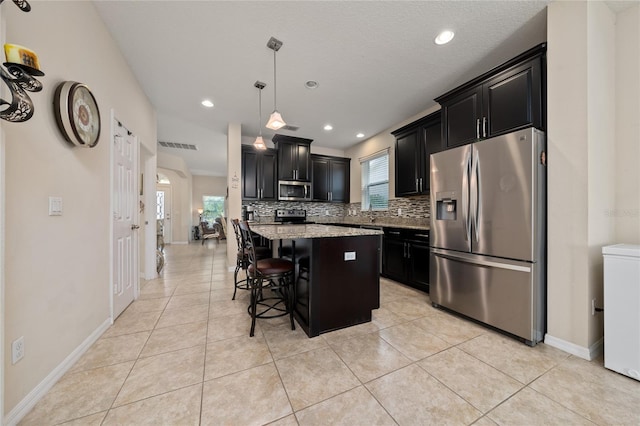 kitchen with a kitchen island, tasteful backsplash, light stone countertops, dark cabinets, and stainless steel appliances