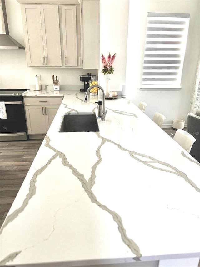 kitchen featuring wall chimney range hood, light stone counters, a sink, electric range oven, and dark wood-type flooring