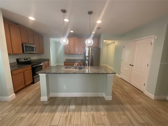 kitchen with dark stone countertops, a sink, light wood-style floors, a kitchen island with sink, and appliances with stainless steel finishes