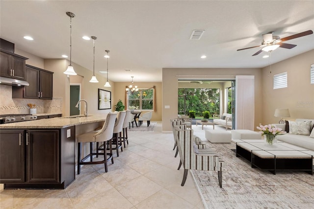 kitchen with a sink, open floor plan, dark brown cabinets, light stone counters, and under cabinet range hood