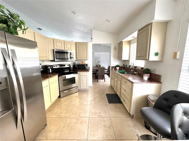 kitchen featuring appliances with stainless steel finishes, a sink, a textured ceiling, cream cabinetry, and vaulted ceiling