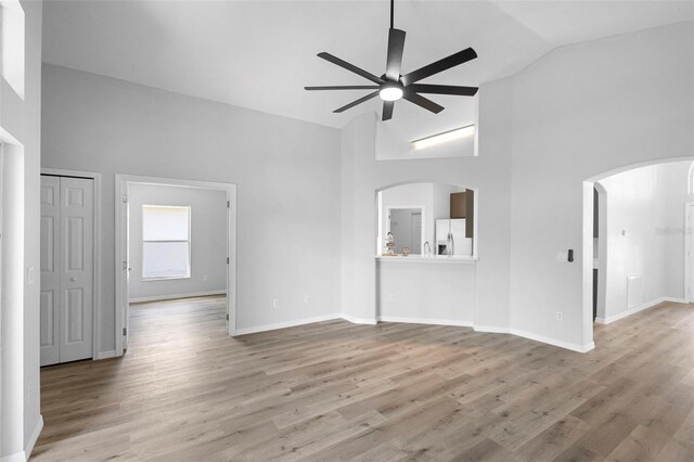 unfurnished living room featuring a ceiling fan, arched walkways, high vaulted ceiling, wood finished floors, and baseboards