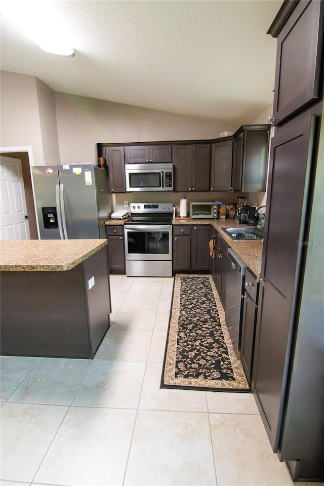 kitchen featuring appliances with stainless steel finishes, a sink, lofted ceiling, dark brown cabinets, and light stone counters
