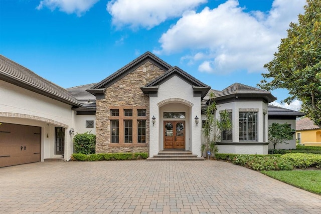 french country inspired facade with stone siding, decorative driveway, stucco siding, and french doors