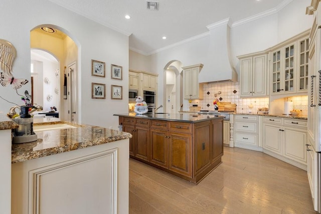 kitchen with cream cabinetry, stainless steel oven, premium range hood, a kitchen island with sink, and crown molding