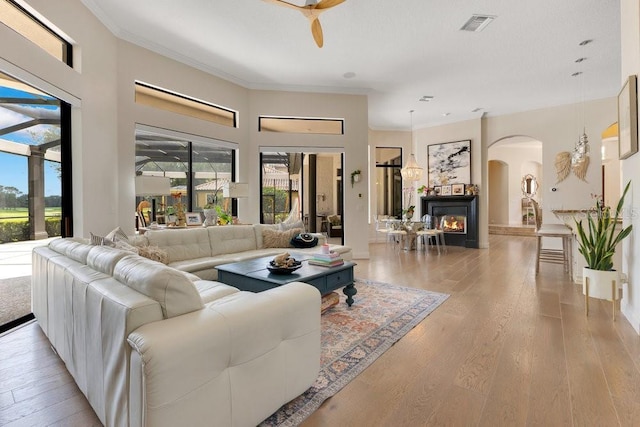 living area featuring arched walkways, a glass covered fireplace, light wood-style flooring, ornamental molding, and a ceiling fan