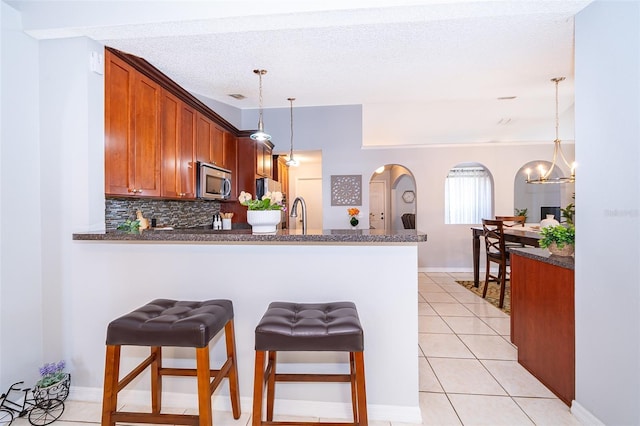 812 Adour Dr, Kissimmee, FL 34759 - kitchen featuring stainless steel microwave, a peninsula, arched walkways, brown cabinetry, and pendant lighting