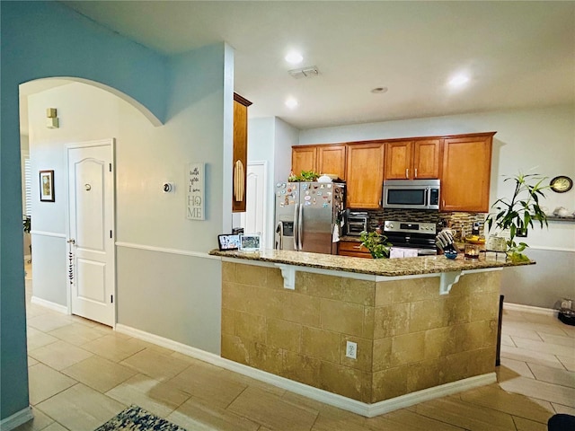 2862 Grasmere View Pkwy, Kissimmee, FL 34746 - kitchen featuring appliances with stainless steel finishes, stone counters, brown cabinets, a peninsula, and arched walkways