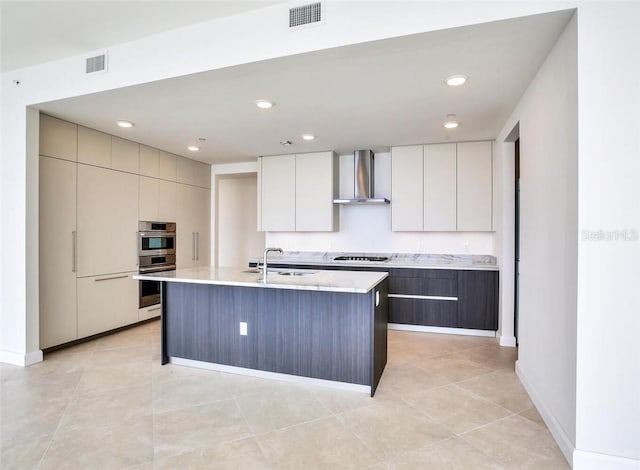 kitchen with stainless steel double oven, gas stovetop, sink, a center island with sink, and wall chimney range hood