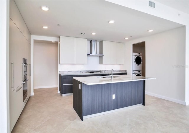 kitchen featuring wall chimney exhaust hood, a center island with sink, gas stovetop, sink, and stacked washer / dryer