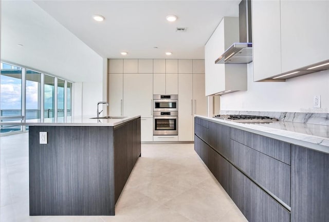 kitchen with gray cabinetry, appliances with stainless steel finishes, sink, wall chimney exhaust hood, and a kitchen island with sink