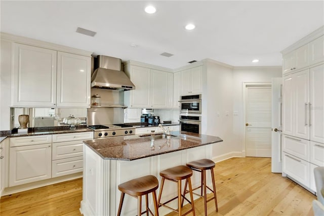 kitchen featuring white cabinetry, a kitchen island with sink, light hardwood / wood-style flooring, dark stone counters, and wall chimney exhaust hood