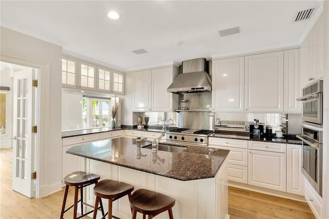 kitchen with an island with sink, wall chimney exhaust hood, light hardwood / wood-style floors, and white cabinetry