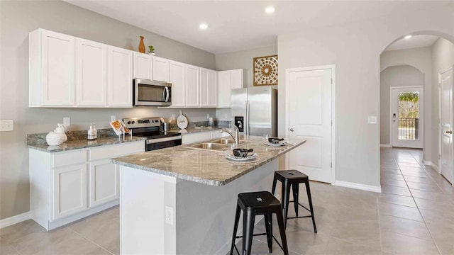 kitchen with an island with sink, white cabinetry, stainless steel appliances, light stone countertops, and sink