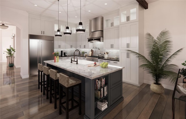 kitchen featuring hanging light fixtures, dark wood-type flooring, a kitchen island with sink, white cabinets, and wall chimney range hood