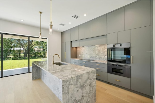 kitchen with sink, light stone counters, black electric cooktop, an island with sink, and gray cabinets