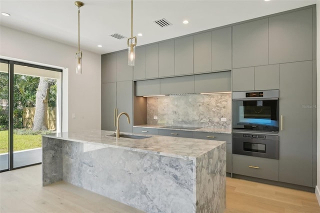 kitchen with sink, gray cabinets, black electric cooktop, an island with sink, and double oven