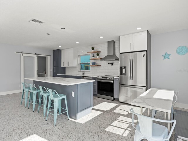 kitchen featuring light stone counters, a barn door, tasteful backsplash, wall chimney range hood, and stainless steel appliances