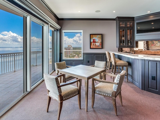 dining room with indoor wet bar, light colored carpet, a water view, ornamental molding, and wainscoting