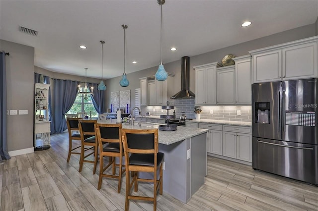 kitchen featuring wall chimney range hood, a breakfast bar area, decorative light fixtures, an island with sink, and stainless steel refrigerator with ice dispenser