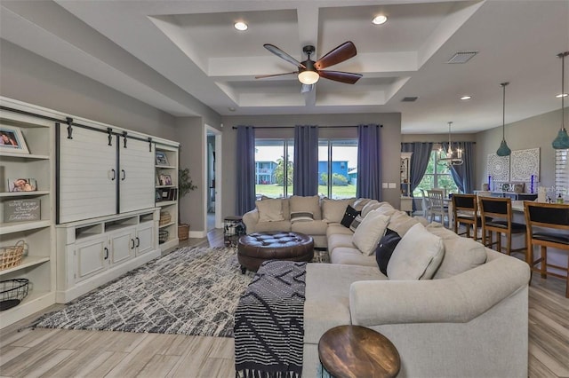 living room with a barn door, ceiling fan with notable chandelier, a raised ceiling, and light hardwood / wood-style flooring