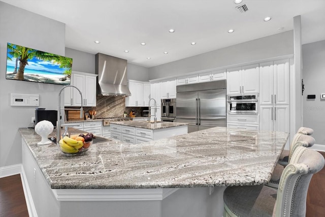 kitchen featuring built in fridge, wall chimney range hood, a large island, light stone countertops, and white cabinets