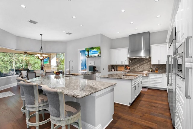 kitchen with pendant lighting, white cabinetry, an island with sink, light stone counters, and wall chimney range hood