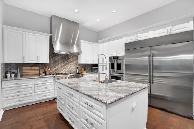 kitchen with white cabinetry, stainless steel appliances, light stone countertops, an island with sink, and wall chimney exhaust hood