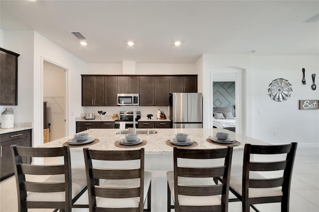 kitchen with stainless steel appliances, a center island with sink, light tile floors, and dark brown cabinetry