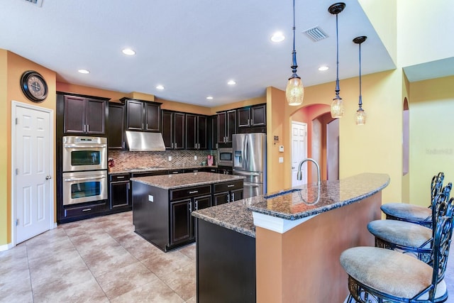 kitchen with a kitchen island with sink, decorative light fixtures, backsplash, appliances with stainless steel finishes, and dark stone counters