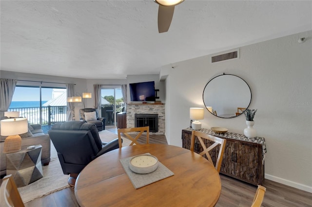 dining space featuring wood-type flooring, ceiling fan, a water view, and a stone fireplace